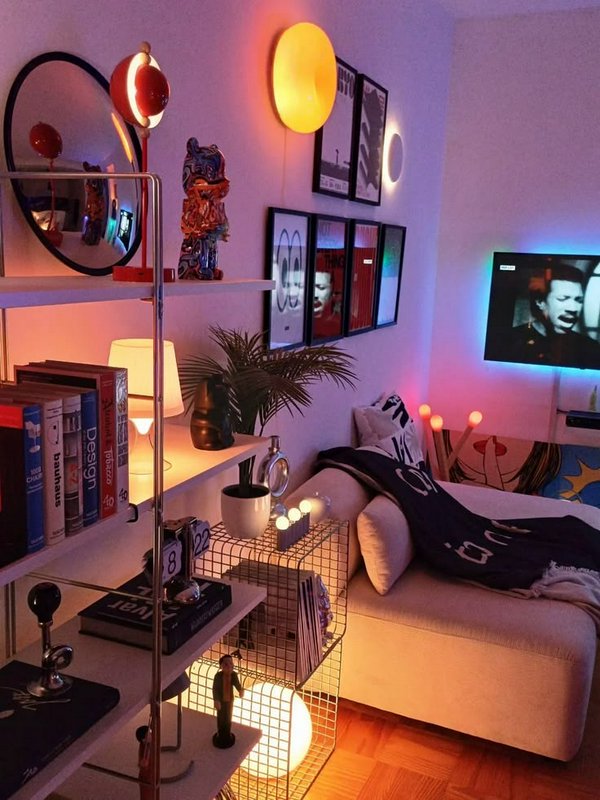 Maximalist cozy apartment nook with warm mood lighting, shelving, framed art, and a plush daybed