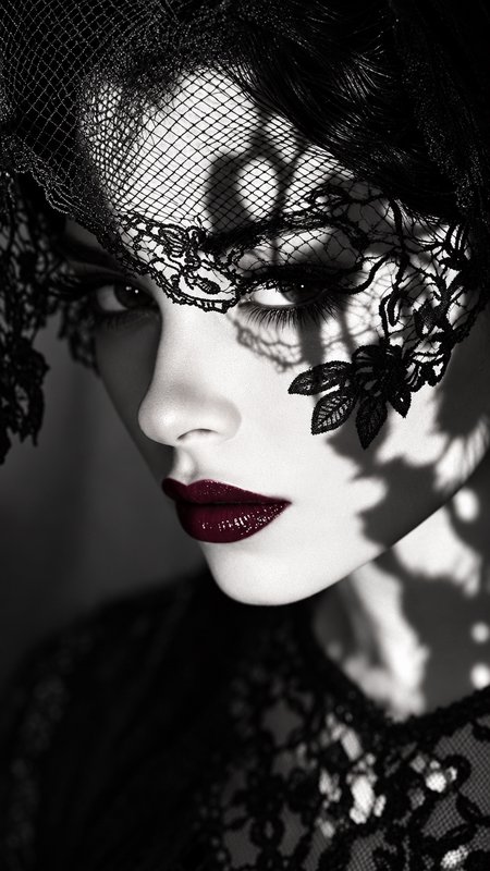 Close-up portrait of a woman in an ornate black lace veil, with bold glossy dark red lipstick against monochrome styling