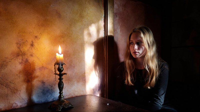 Young woman with long blonde hair sits in a dim room beside a lit candle on an ornate candlestick.