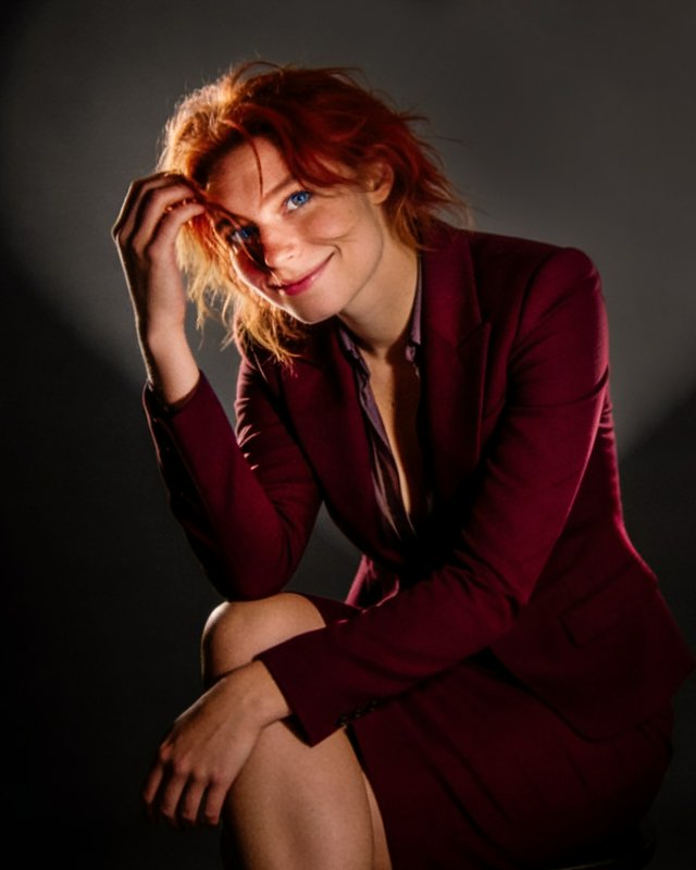 Red-haired woman in a burgundy blazer seated, touching her hair in a moody studio portrait.