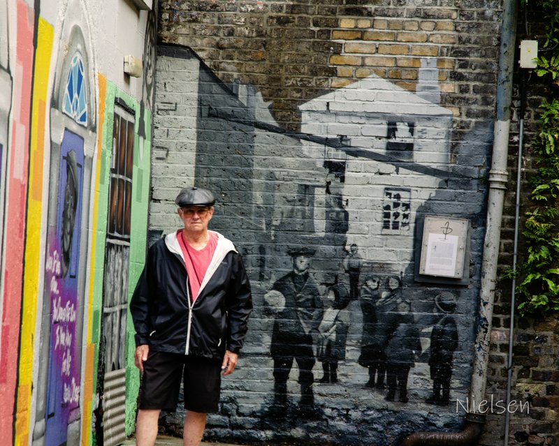 Senior man in a flat cap stands before a historic street scene mural on a weathered brick alley wall.