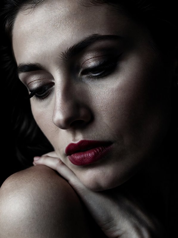 [Close-up low-key portrait of a pensive woman with bold red lipstick, eyes cast downward, resting her hand on her shoulder...