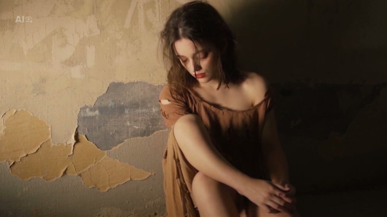 Pensive young woman in a tattered brown dress sits with head bowed against a peeling weathered wa...