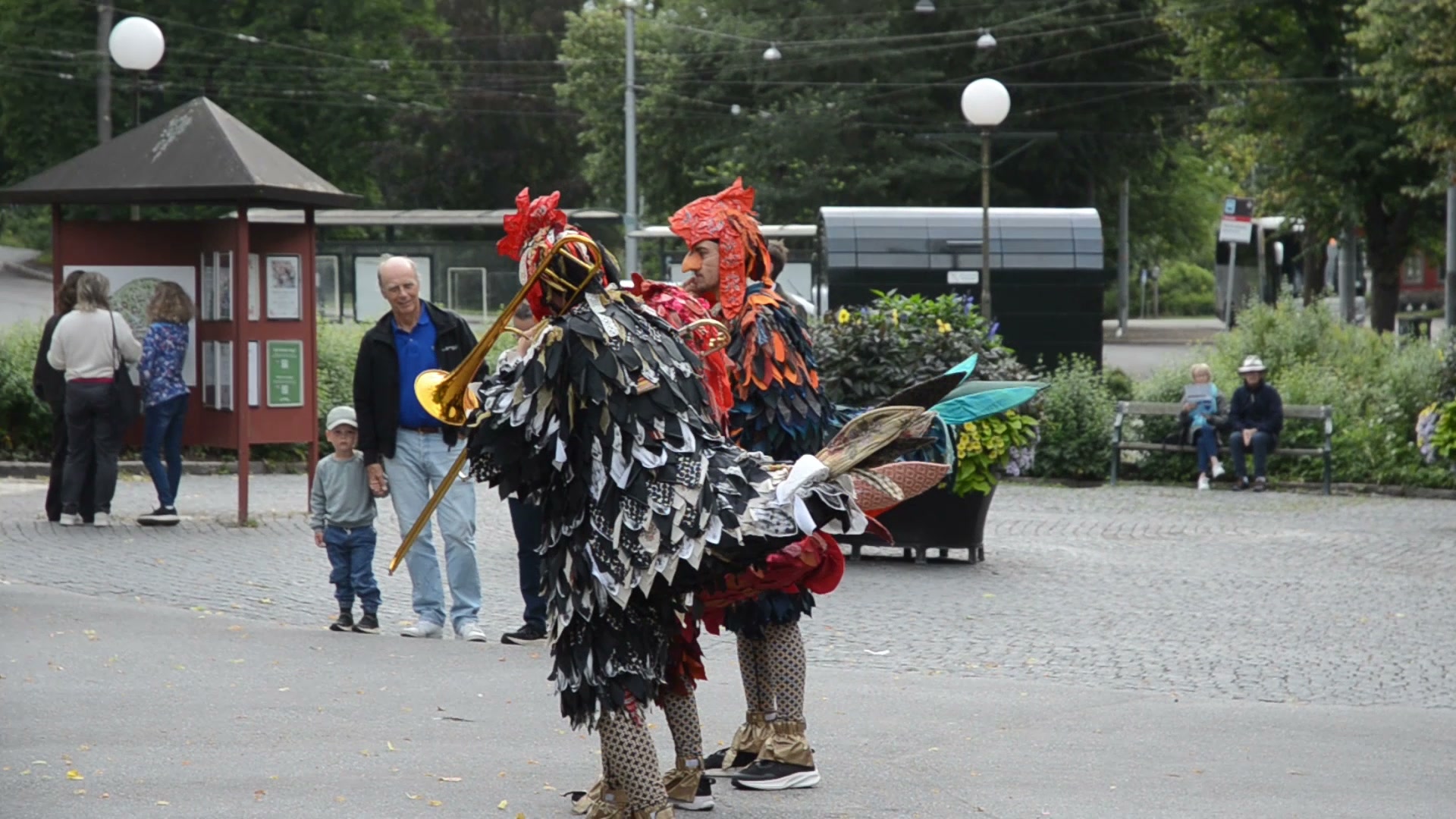 Street performers in elaborate rooster costumes play brass instruments for onlookers in a public ...