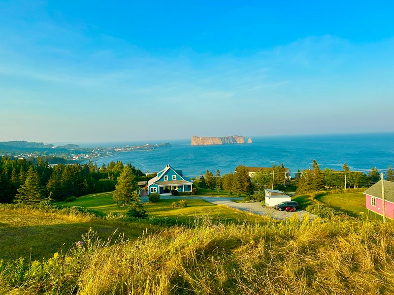 Coastal hillside with a blue house, pine trees, and the sea in the distance under a clear blue sky.