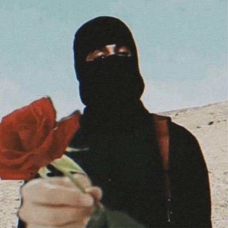 Person in a black niqab offering a red rose in a desert.