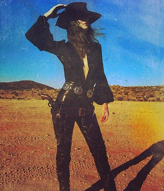 Glamorous vintage styled cowgirl in black western wear, wide hat, and holstered revolver stands i...