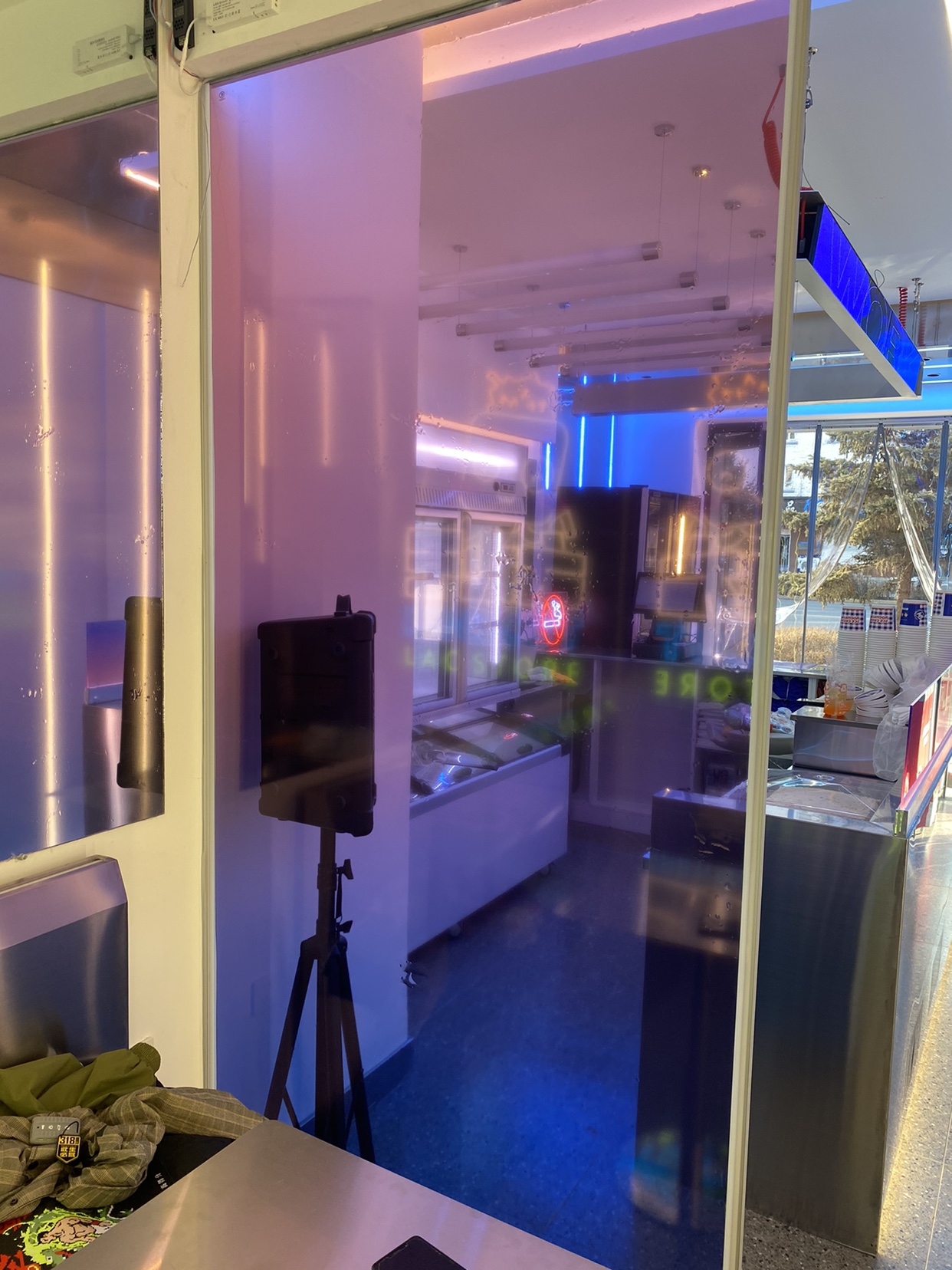 Neon-lit shop interior with glass doors, display case, and a tripod-mounted speaker.