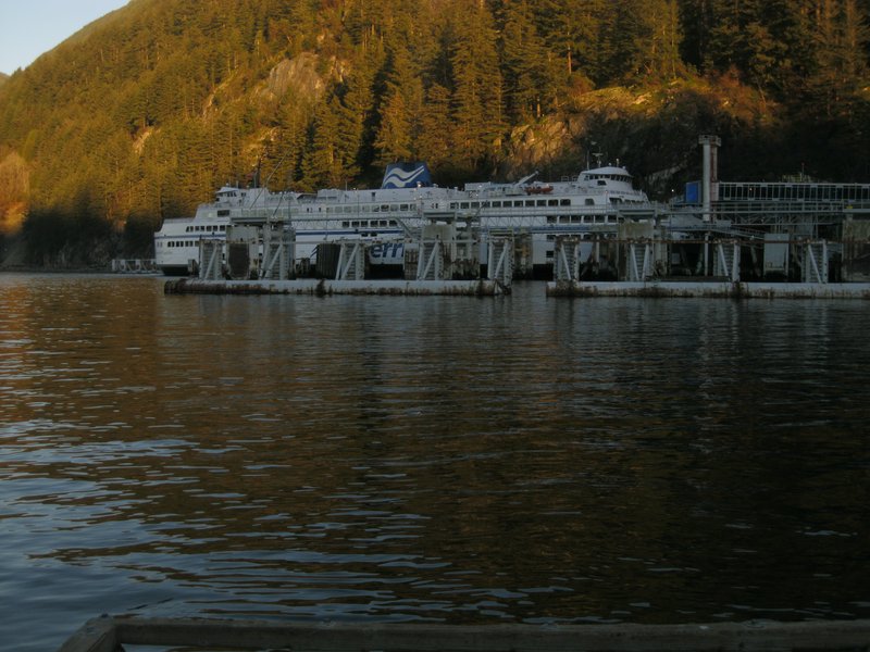 Large white ferry docked at waterfront terminal, calm inlet water, sunlit evergreen forested coas...