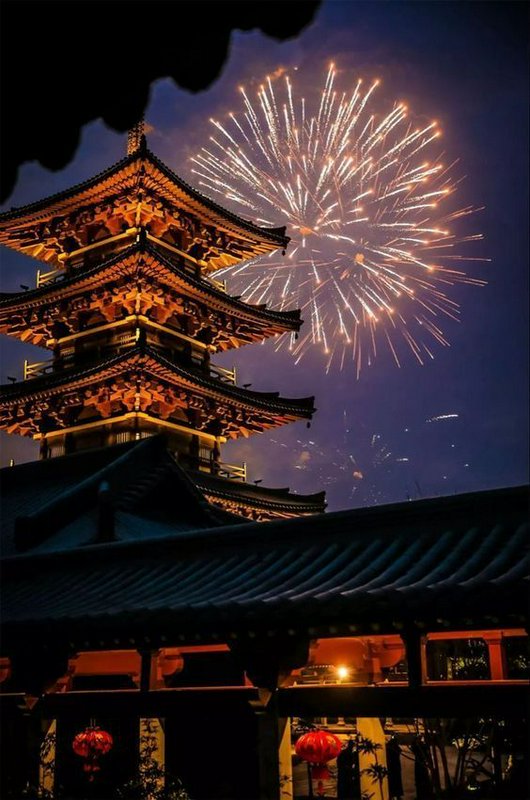 Night view of a multi-tiered Japanese pagoda with fireworks bursting in the sky behind it.