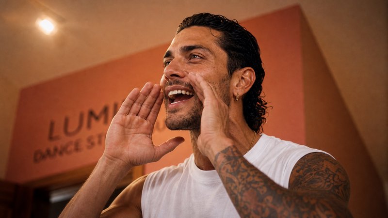 Candid shot of a smiling tattooed man cupping his hands to shout inside a warm-toned dance studio