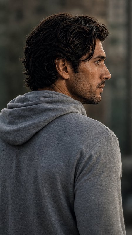 Profile view of a rugged stubbled man with wavy dark hair, wearing a gray hoodie, gazing right ag...