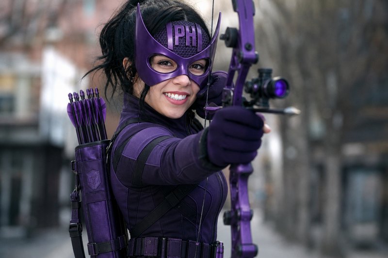 Female archer in a purple superhero cosplay aiming a bow in an urban street.