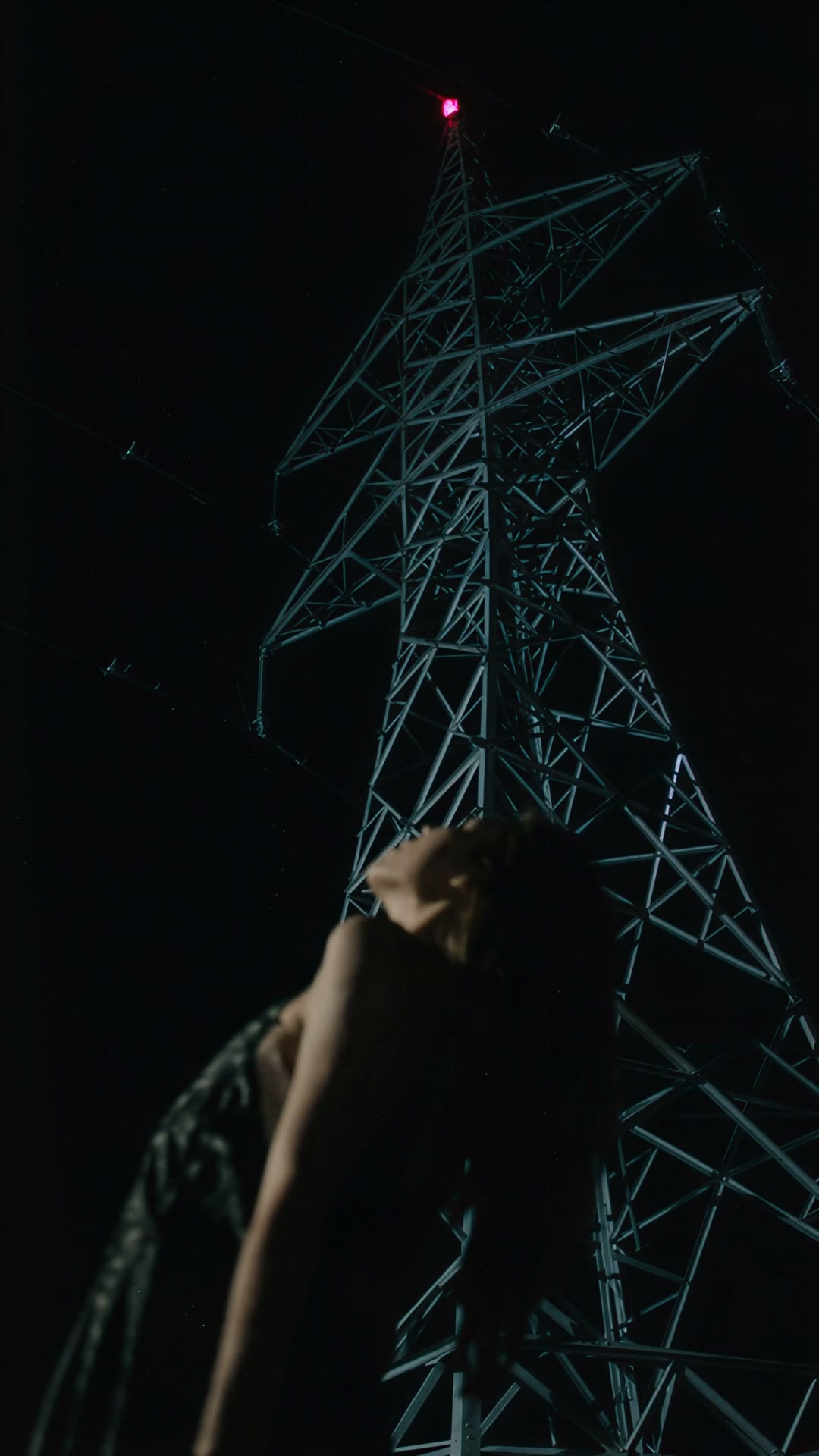 Silhouetted woman leans back looking up at a lit steel electricity pylon under a dark night sky w...