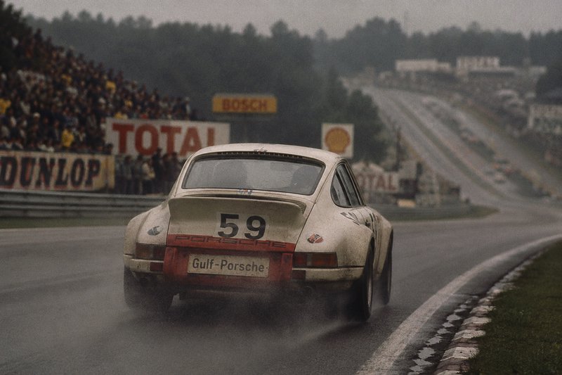 Rear view of #59 Gulf-Porsche 911 race car driving on a wet foggy track, with tire spray kicking ...