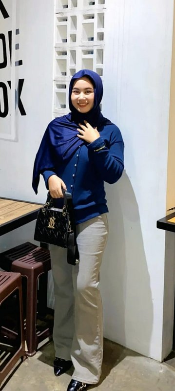 Woman in a blue hijab and sweater posing in a cafe with a black quilted handbag.
