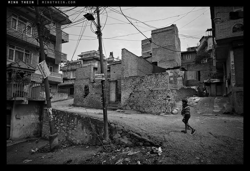 A lone boy walking through a ruined urban alley with damaged brick buildings and tangled wires.