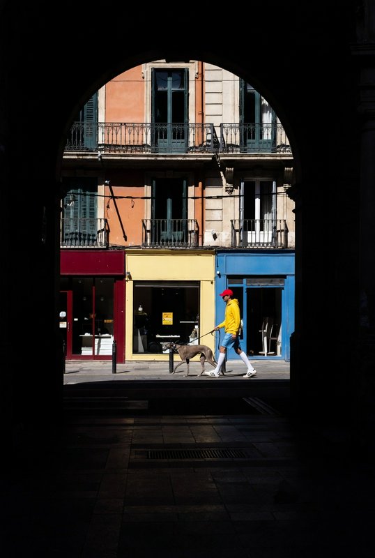 Person in a yellow jacket and red cap walks a dog through a dark archway toward a colorful storef...