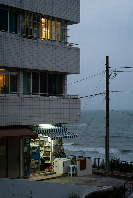 Coastal tiled apartment building with glowing ground-floor corner store overlooking choppy overca...