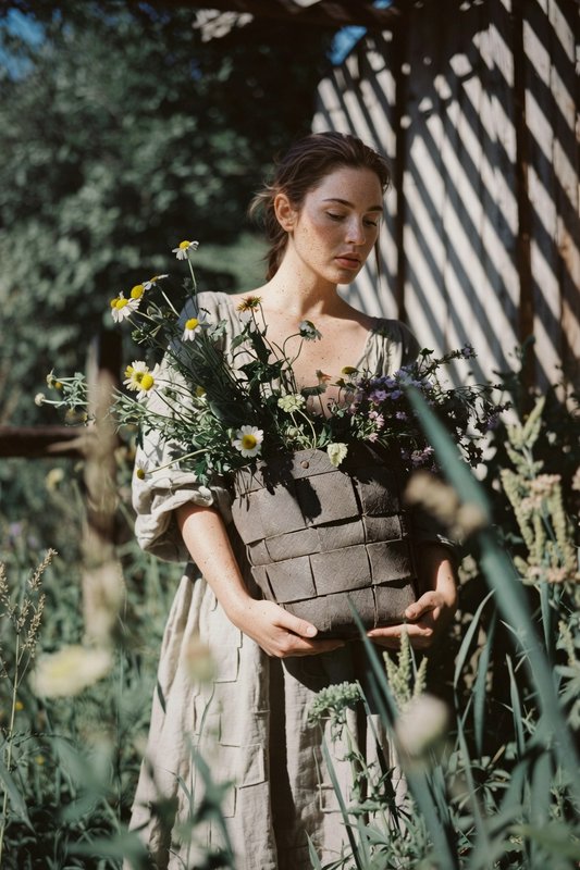 Soft portrait of a woman in a linen dress holding a basket of fresh wildflowers in a sun dappled ...