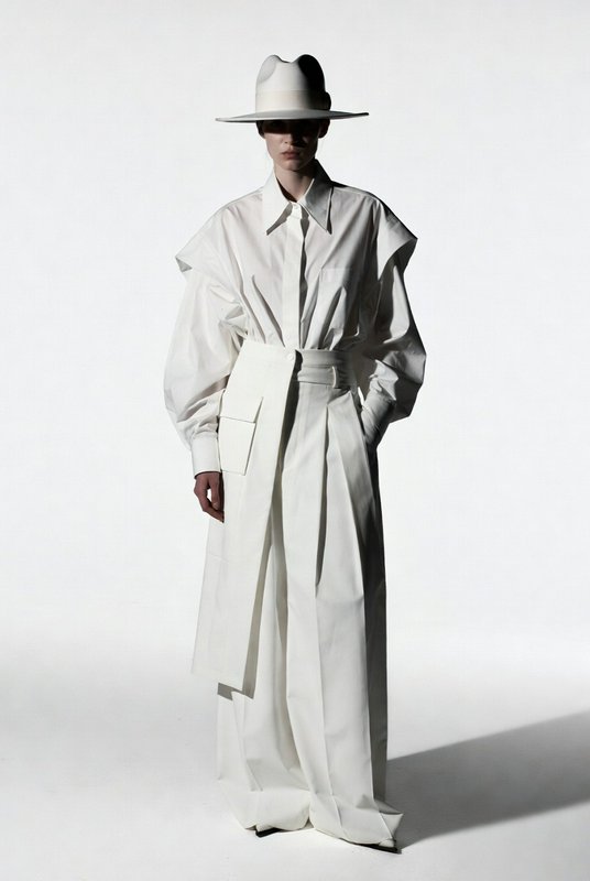 Model in all-white avant-garde oversized outfit and wide brim hat poses against plain white studi...
