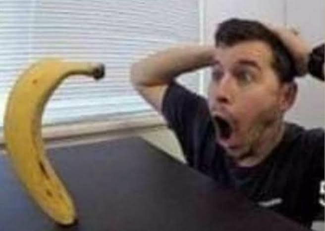Man with shocked expression at a curved banana on a table in an office setting.