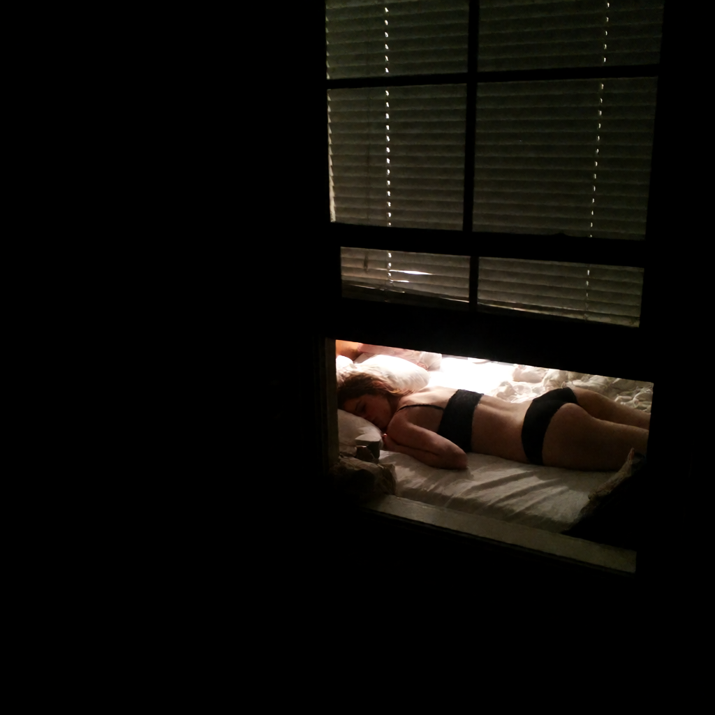 View through a dark window frame of a woman in black lingerie sleeping face down on a sunlit rumpled bed.