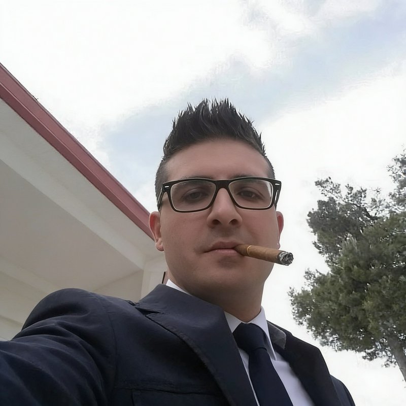 Low angle outdoor selfie of a suited man with glasses and spiky hair smoking a lit cigar