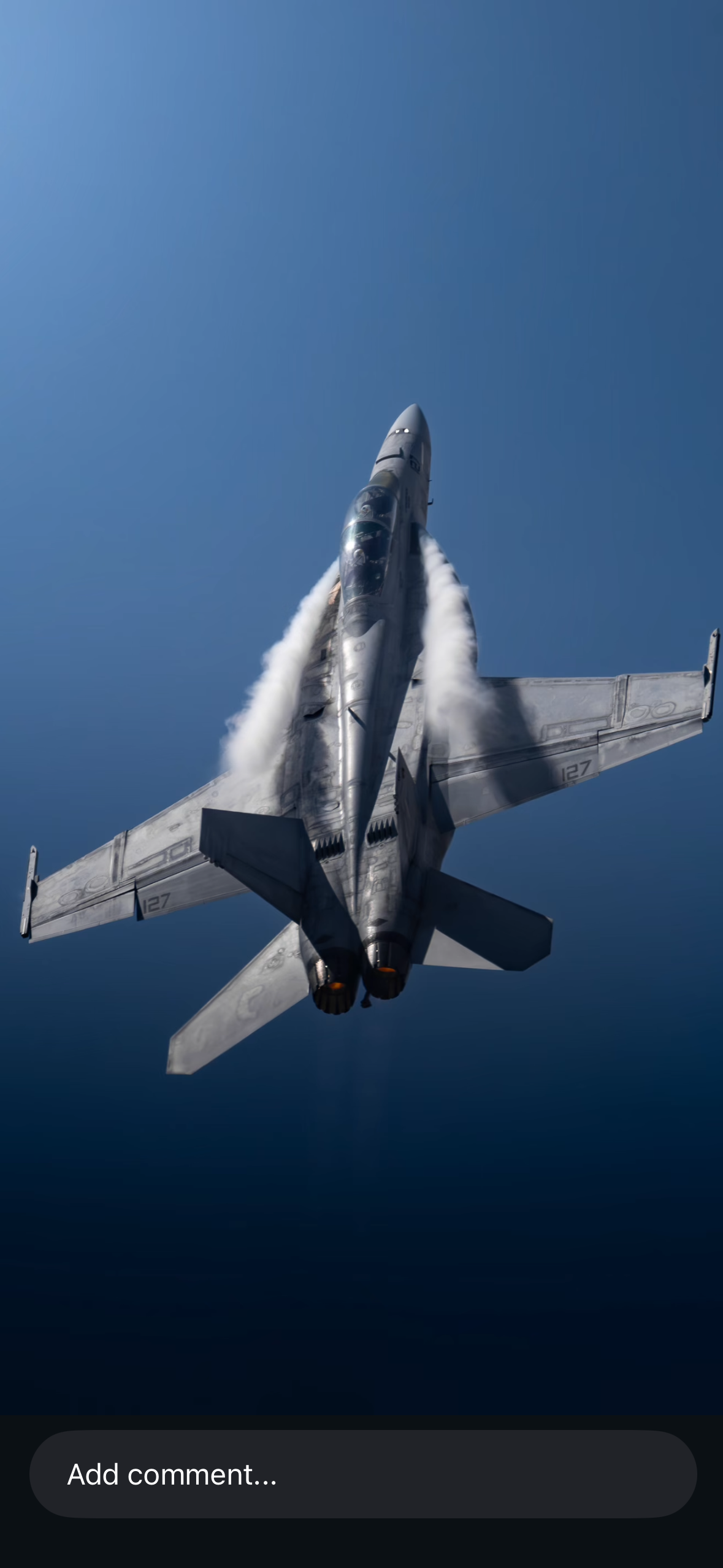 A fighter jet climbs skyward with afterburners, viewed from behind against a deep blue sky.