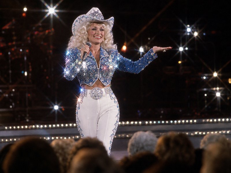 Dolly Parton performs on a lit concert stage in a sparkly rhinestone western cowboy outfit, holding a microphone and