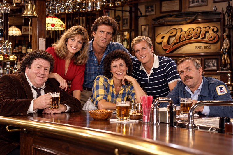 Cheers sitcom main cast smiling while posing behind the show's iconic wooden pub bar