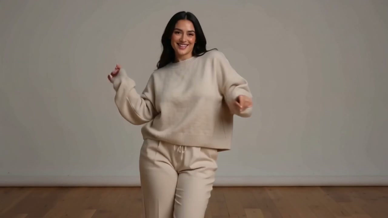 Smiling woman in a beige loungewear set playfully poses in a plain neutral studio with hardwood f...