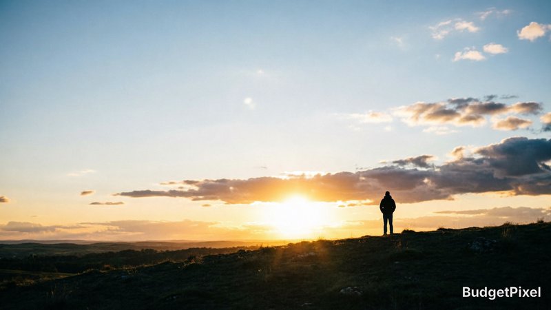 [Silhouetted lone person standing on a grassy hilltop watching a golden sunset over rolling rural countryside]