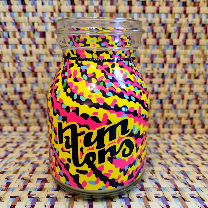 Bright neon patterned glass jar with stylized lettering sits on a multicolor woven mat