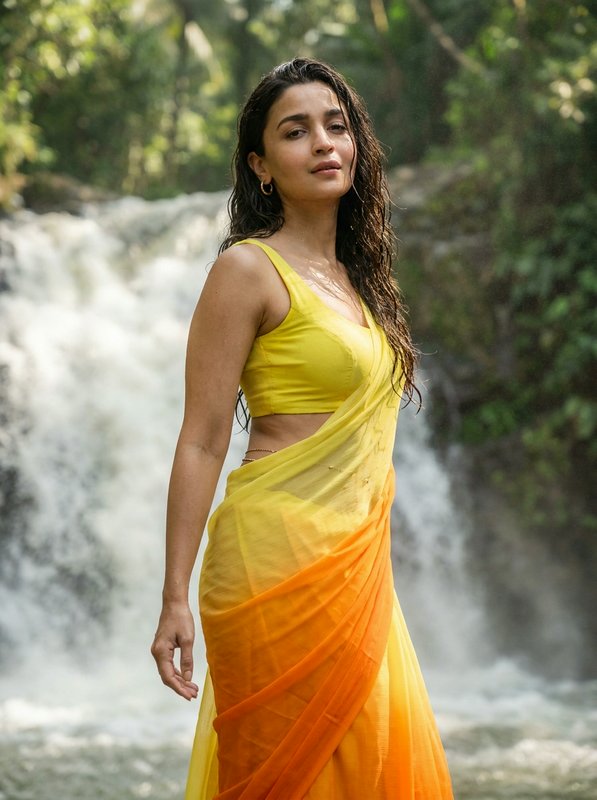 Alia Bhatt in a yellow-to-orange gradient saree, standing before a lush rainforest waterfall with...