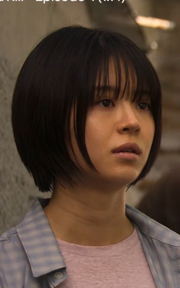 Woman with a short black bob and bangs wearing a pink top and blue plaid shirt in an urban subway setting.