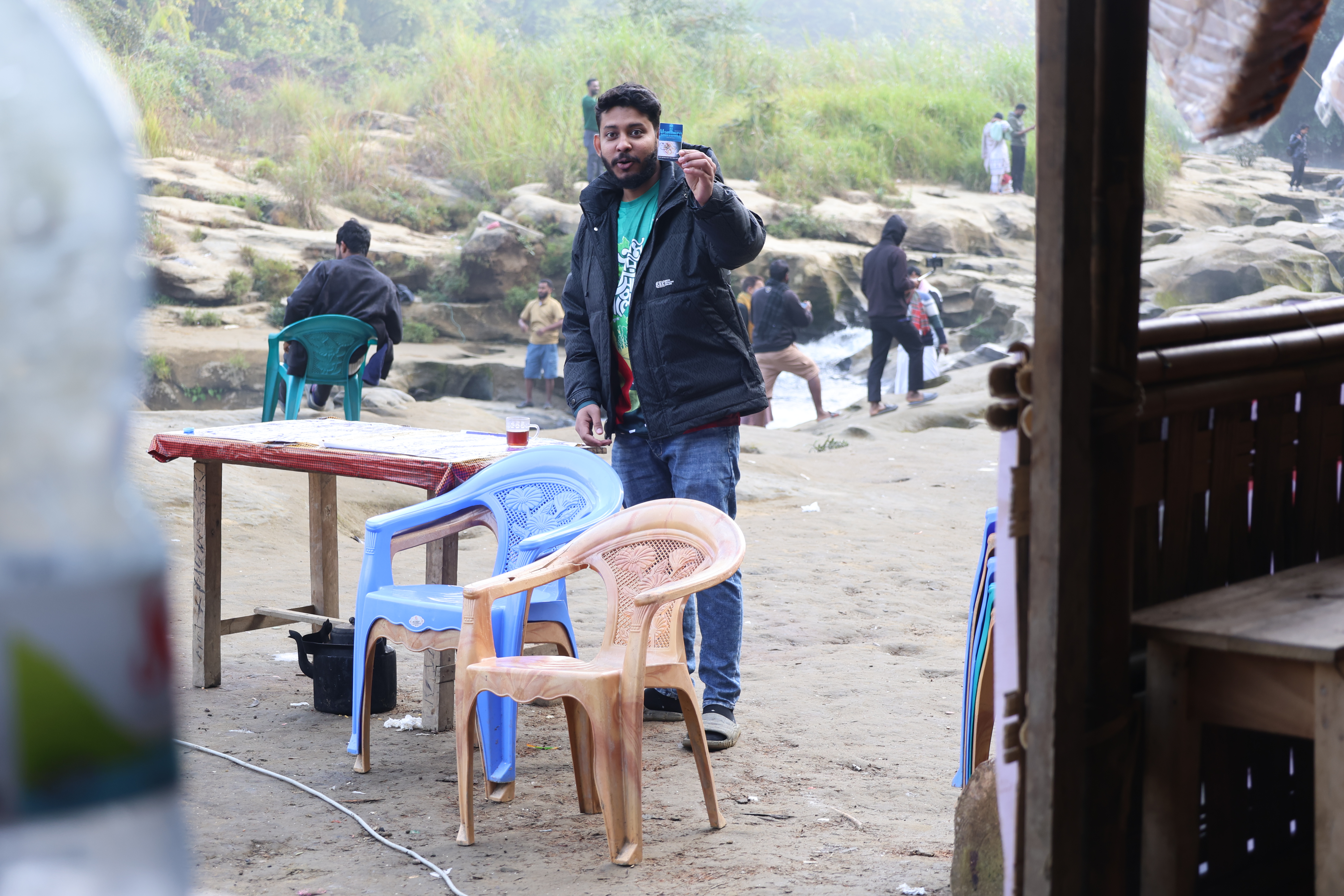 man taking a selfie on a sandy riverside with colorful plastic chairs and a wooden table