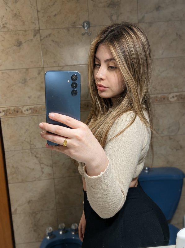 Young woman with highlighted blonde hair takes a mirror selfie with a blue smartphone in a tiled ...