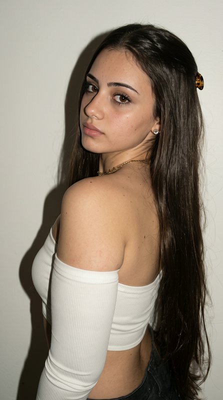 Young woman with long dark hair poses looking over her shoulder, wearing an off-shoulder white to...