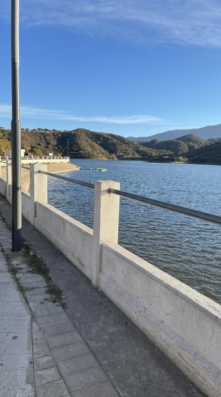 Paved waterfront walkway with concrete railing overlooking calm reservoir, rolling forested hills under bright clear blue ...