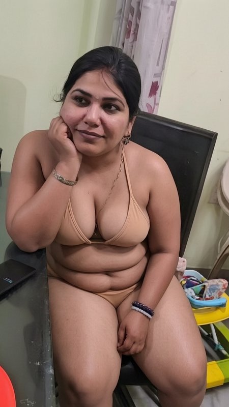 Curvy South Asian woman in a beige bikini rests her chin on her hand while seated indoors at home