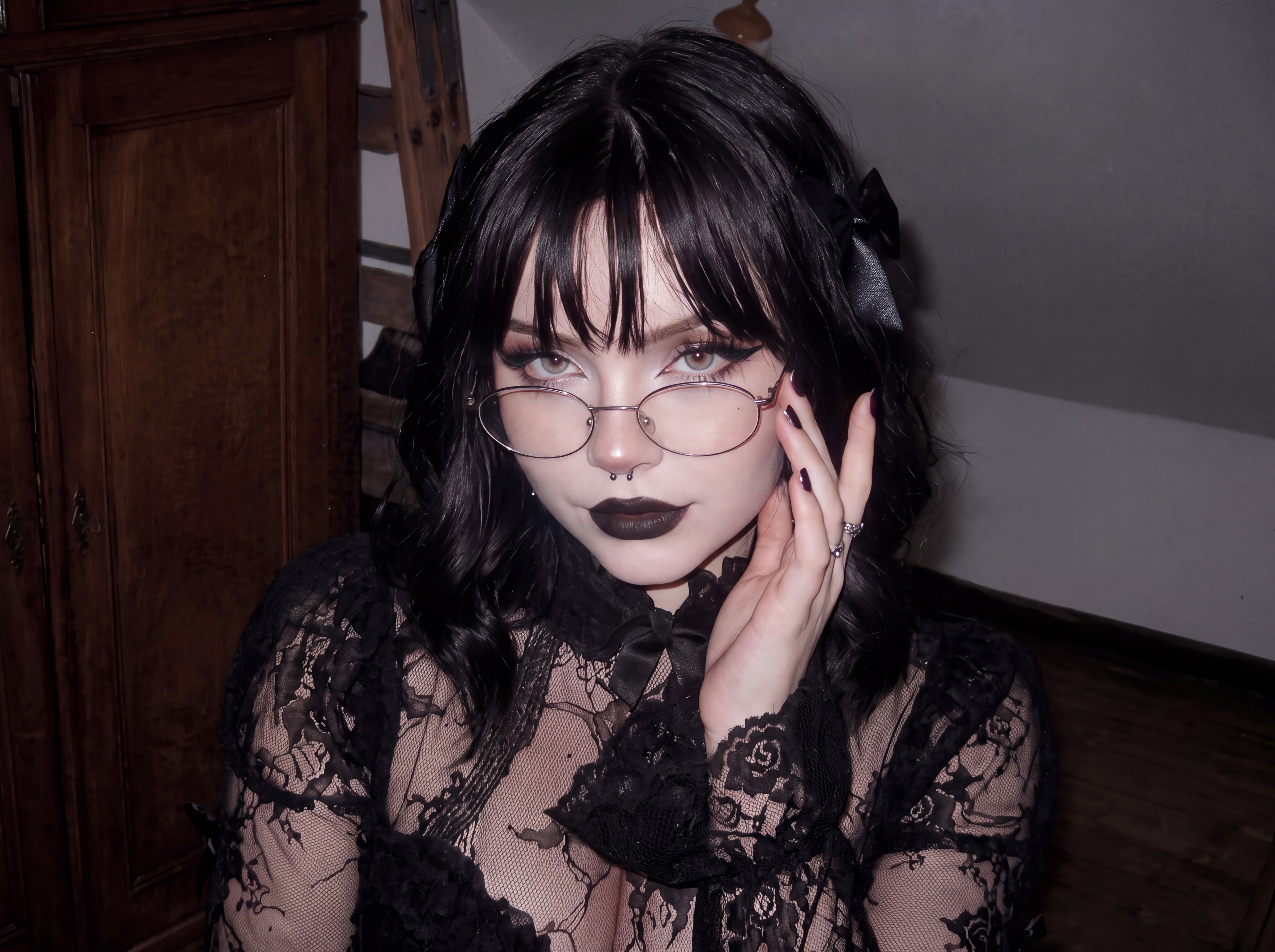 Gothic young woman with round glasses rests her hand on her cheek while wearing a black lace top