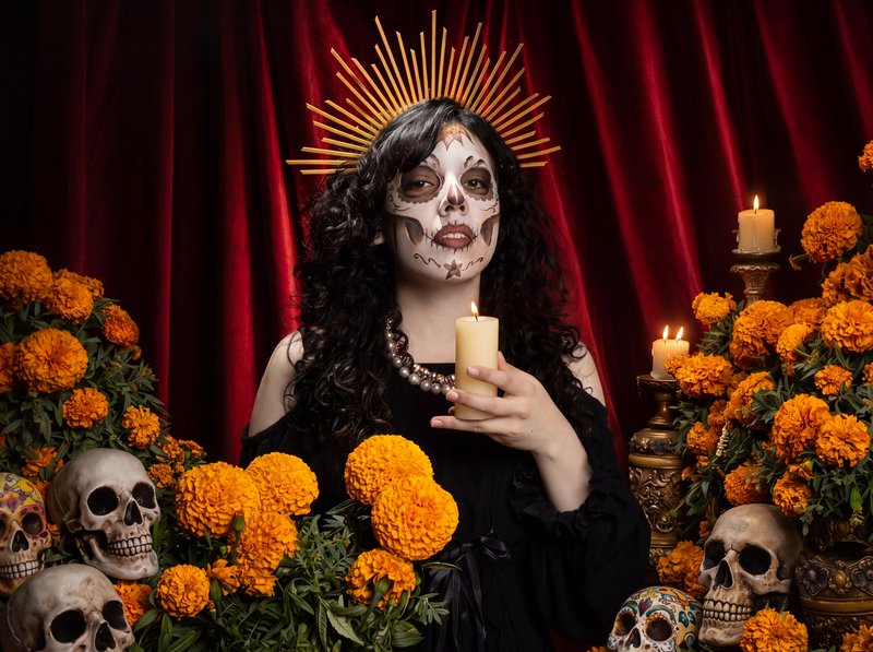 Woman wearing Catrina sugar skull makeup and golden halo holds a candle surrounded by marigolds f...
