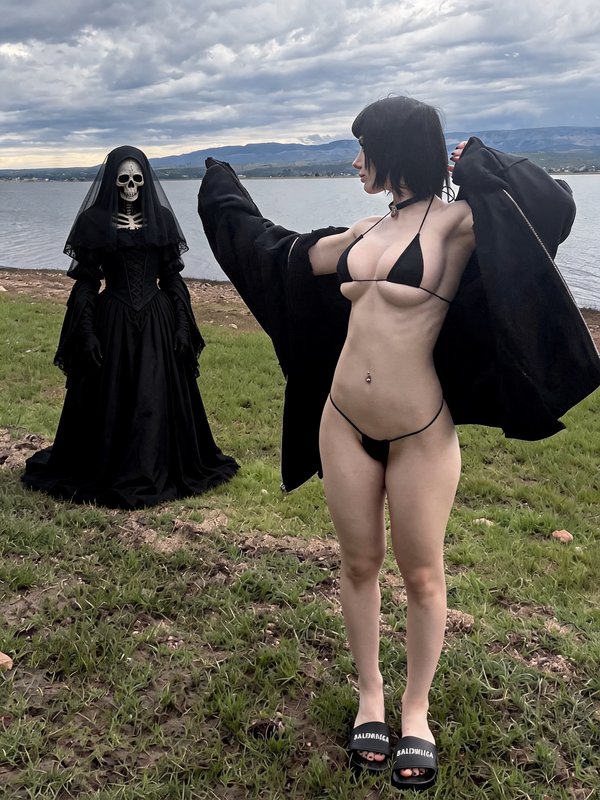Cosplayer in a black micro bikini stands beside a veiled skeleton grim reaper figure on a grassy ...