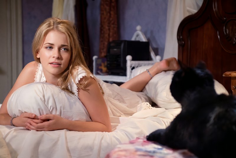 Young blonde woman lying on a bed gazing at a black cat in a cozy bedroom