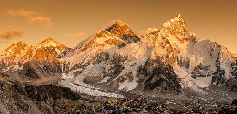 Snowy alpine peaks bathed in orange sunrise above a colorful base camp of tents.