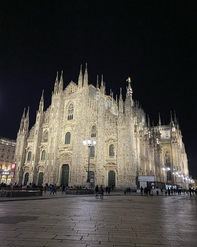 Illuminated Gothic Milan Duomo cathedral stands over Piazza del Duomo at night, with visitors wal...