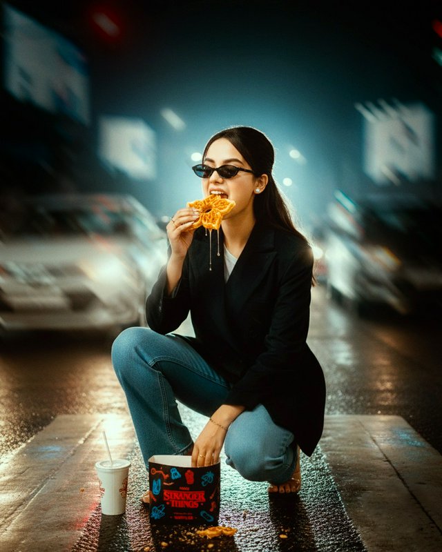 Woman in blazer and jeans kneels on a wet city street at night, eating fried chicken from a Stranger Things box.