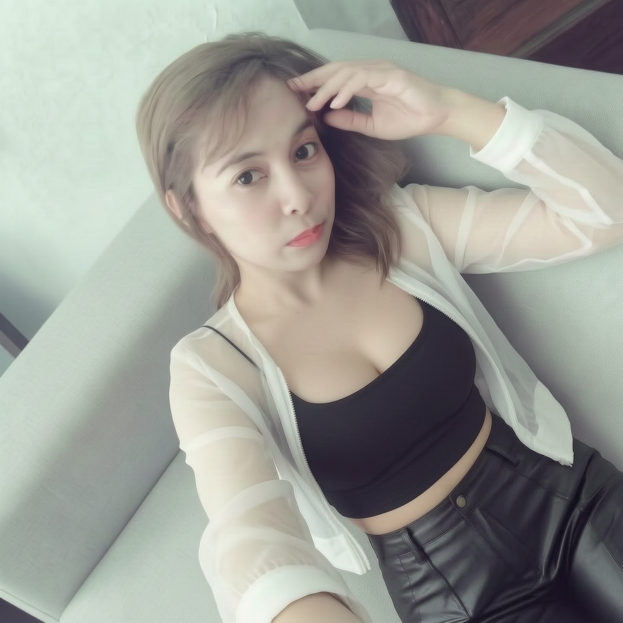 Young woman on a light sofa wearing a black crop top and sheer white jacket, posing for a selfie.