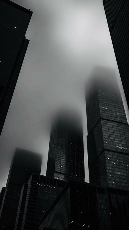 "Low-angle monochrome view of fog-obscured downtown glass skyscrapers, their upper towers disappe...